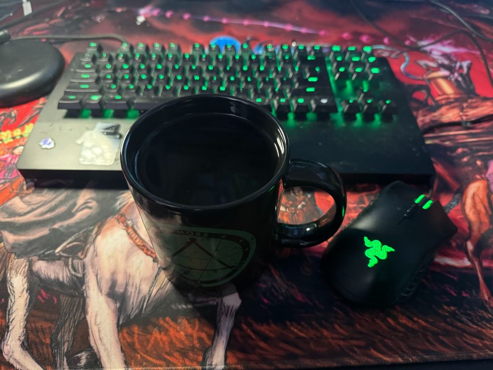 A huge coffee-filled Delta Green mug next to a keyboard and mouse, both with green highlights, all on a large Haunted West mouse pad.