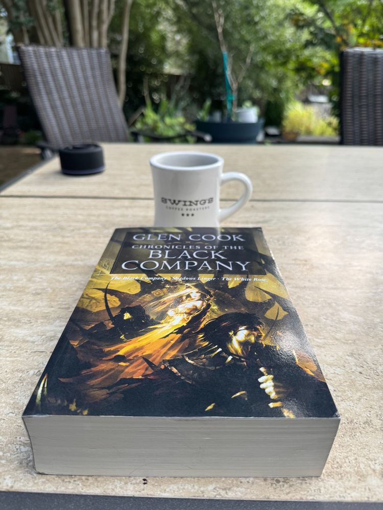 The Chronicles of the Black Company book on a patio table with a Swings Coffee mug slightly behind it. Two patio chairs hazy in the distance. 