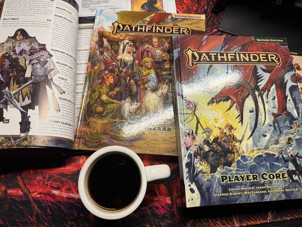 On Chris's desk is a filled coffee cup, Pathfinder Player Core, Pathfinder 2E Grand Bazaar, Pathfinder Dark Archive opened to the image of the Thaumaturge all on a large Haunted West mouse pad showing a demon train. 