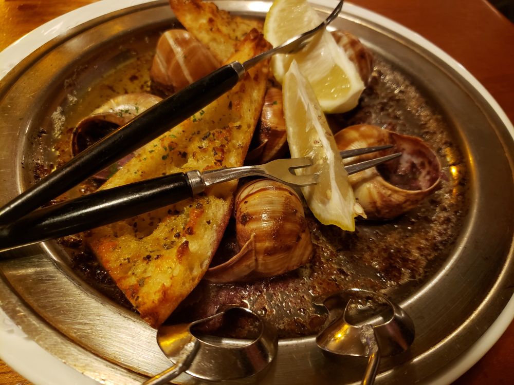 Escargot Bourguignonne - 6 snails in garlic butter on a metal plate with indentation for each snail, with garlic toast and lemon wedges, with special small forks and tongs