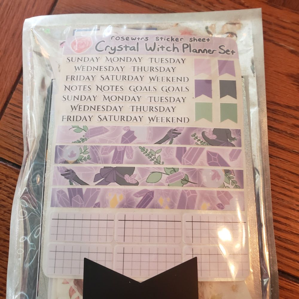 A plastic and foil package of stickers and washi/PET tape. The visible sheet reads rosewtrs sticker sheet Crystal Witch Planner Set. Text stickers included on the upper left read Sunday, Monday, Tuesday, Wednesday, Thursday, Friday, Saturday, Weekend, Notes, Notes, Goals, Goals and then repeat the days of the week. To their immediate right are a series of six flags in shades of purple and green. Beneath these are four strips of crystal and plant themed washi tape in the purple and green colors. Beneath those are six small four by eight grid stickers with a pale lavender back ground. 

There are hints at additional sticker sheets beneath, but they are obscured by the black flag tail logo of the branded sticker club package that was used to contain them. 