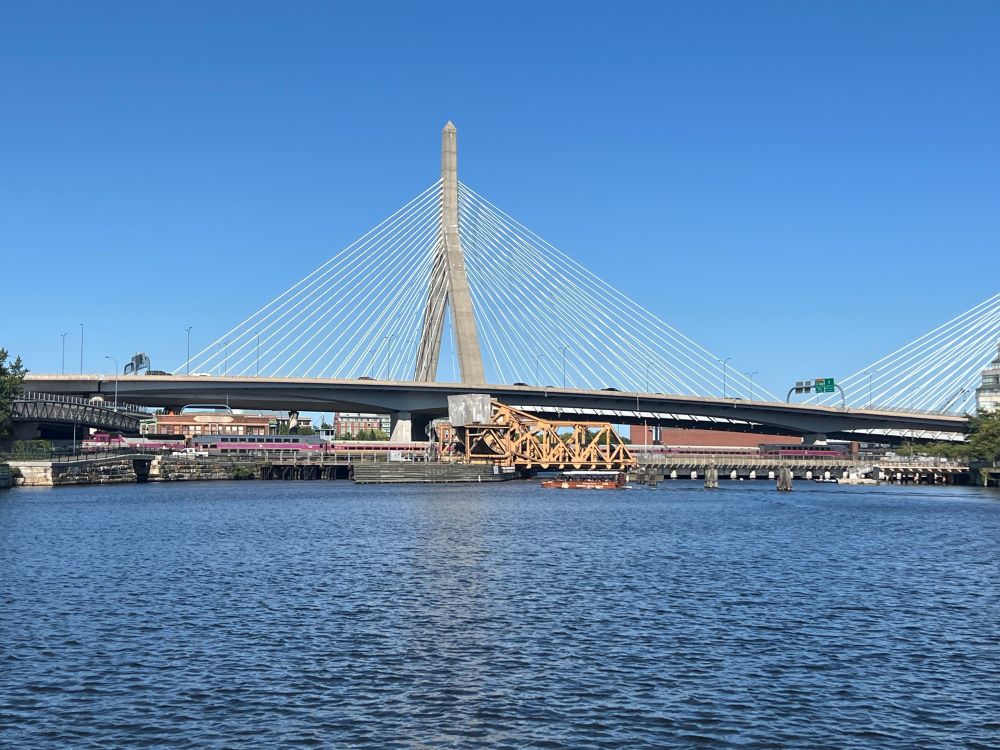 A brown Duck Boat glides through the Charles River on a sunny day. In the background, an MBTA commuter rail train bound for Rockport crosses Draw One as the Leonard P. Zakim Bunker Hill Memorial Bridge looms behind it. 