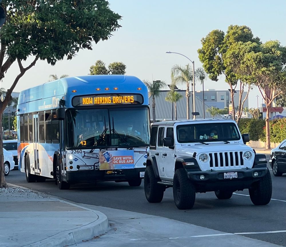Photo of Orange County Transit Authority bus with the head sign saying, “Now Hiring Drivers.” The bus is a New Flyer CNG with a mostly blue livery. 