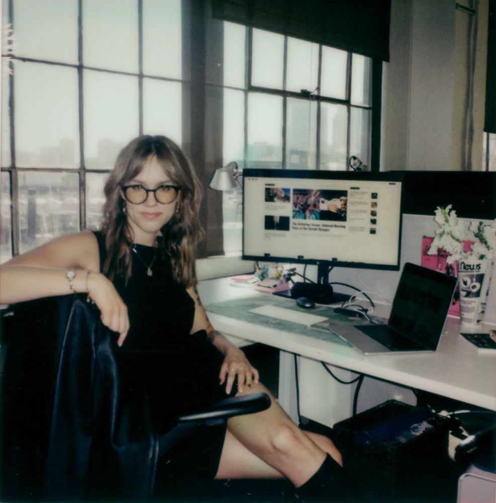 a film photo of me sitting at my desk chair in all black, leaning over the chair slightly with one arm
