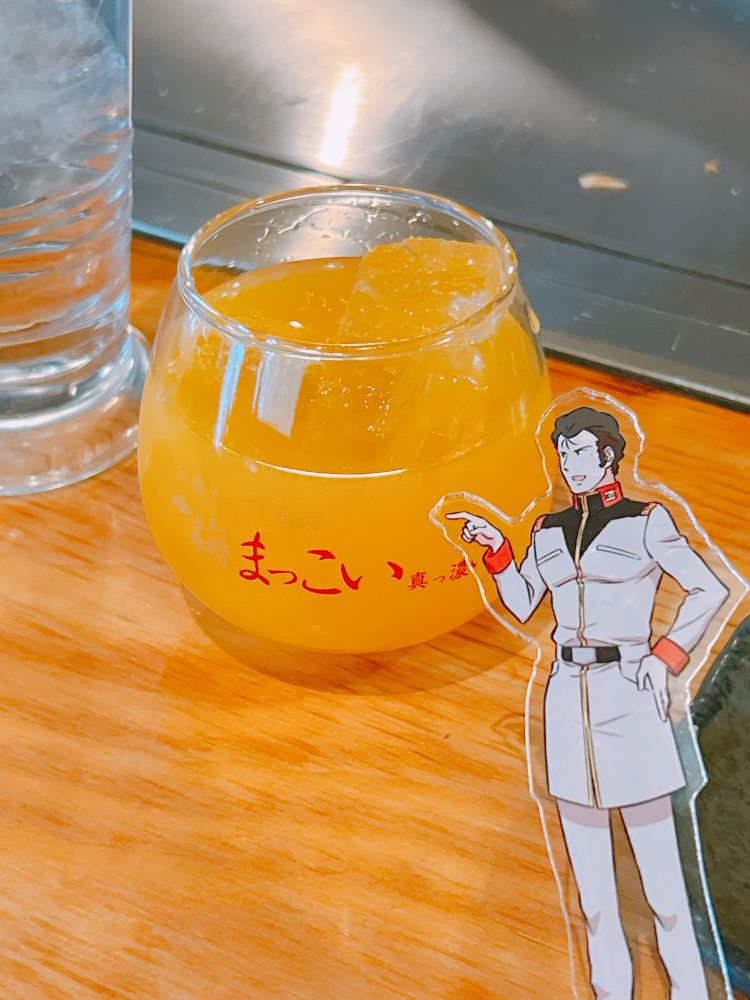 グラスに入ったみかん酒ロック。それを指差すブライトのアクスタ。端にチェイサーの水もある。