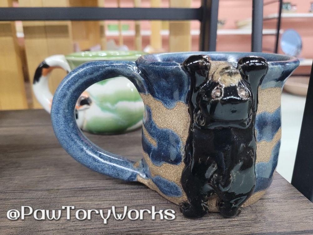 A wavy blue mug on a shelf, featuring a cute sculpted black bear "hanging" from the mug rim by its front paws. 