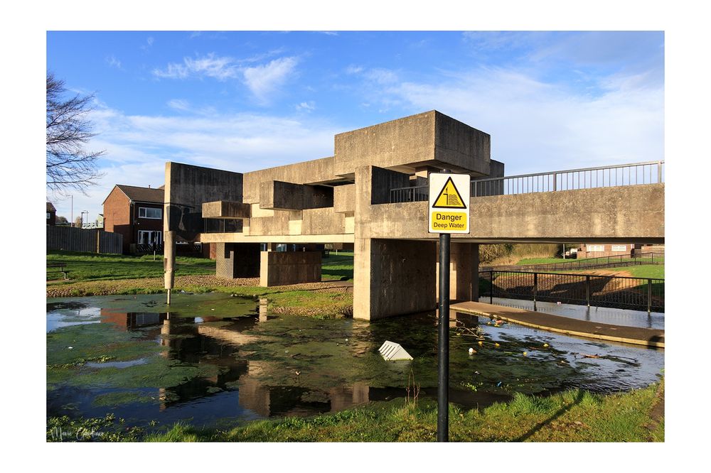 the pavilion from the outside: concrete blocky shapes sitting above water full of trash and algae with a 'danger deep water sign' in the front