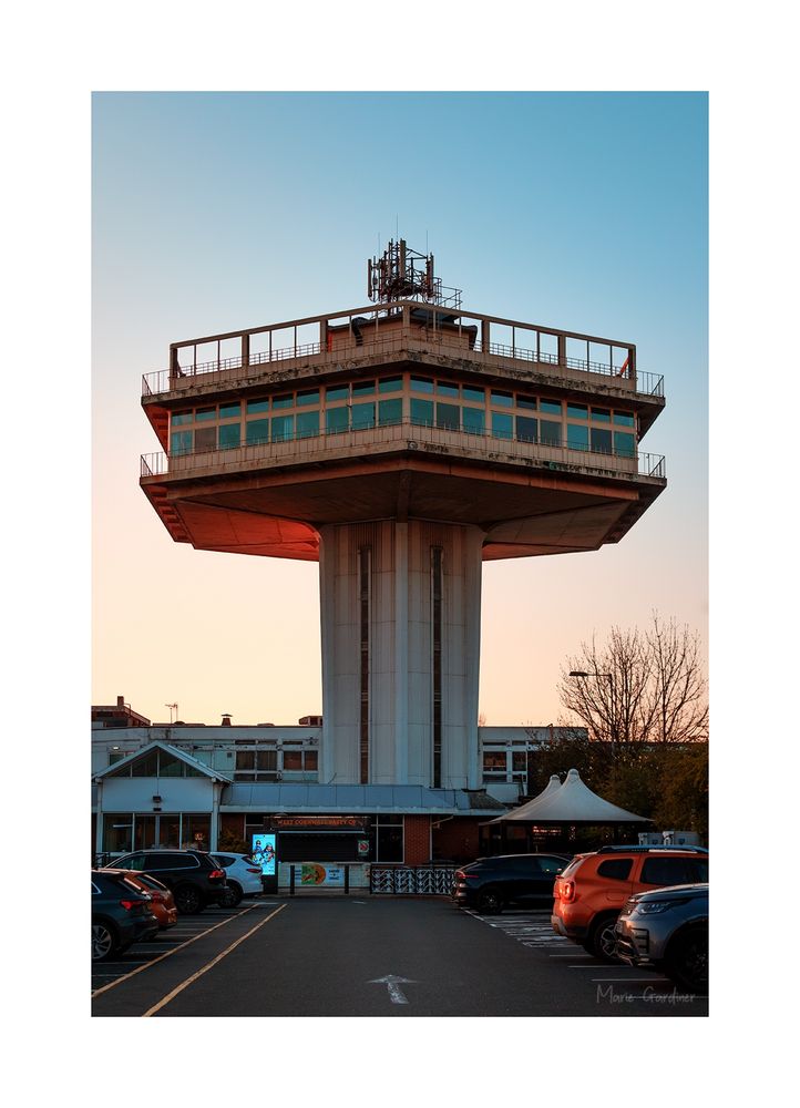 the pennine tower at forton services, an air traffic control tower shape of concrete, with shops and food places below it. Cars can be seen parked on either side of the bottom of the image