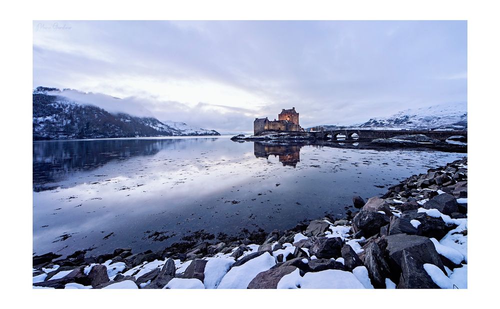 a castle (Eilean Donan) with its bridge leading to it, in the middle of a loch. It's winter and there's a dusting of snow. The colouring is a cold pale purple as the last of the light goes.
