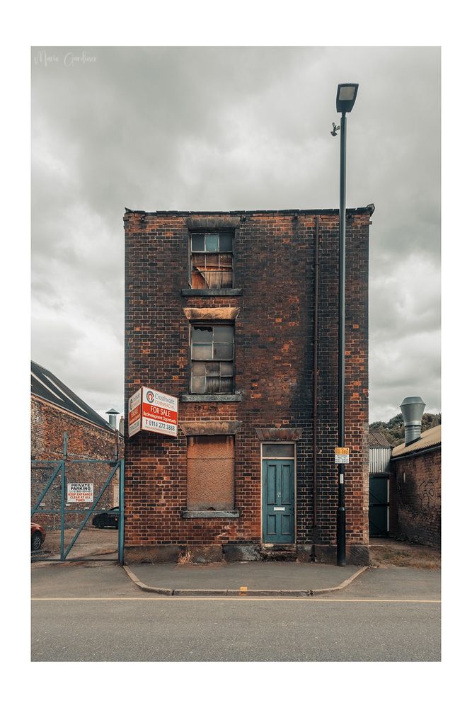a detached three story building in poor shape. The bricks are burnt and the windows are either smashed or boarded up. There's a for sale sign on the lower left third of the building. On the right there's a lamp post with a pigeon that looks a bit fake but isn't, flying past it