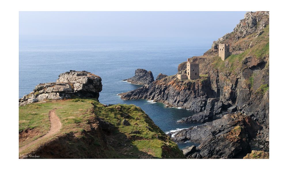the remnants of a tin mine to the right, on a cliff, with a path leading across another smaller cliff to the left, and the sea visible with lots of rocks