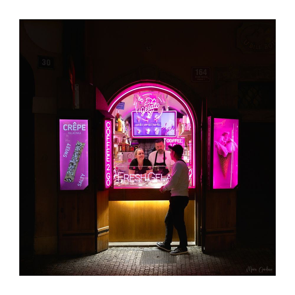 an arch shop hatch selling fresh gelato, ringed in a pink neon light with many other pink themed lit advertisements around it. A person is standing mostly facing away from us, leaning into the hatch and talking to two people behind the counter