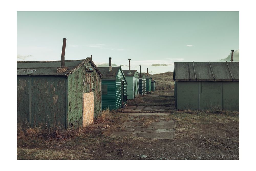 green wooden fishing huts with metal chimneys, standing in rows, the one closest to us is boarded up