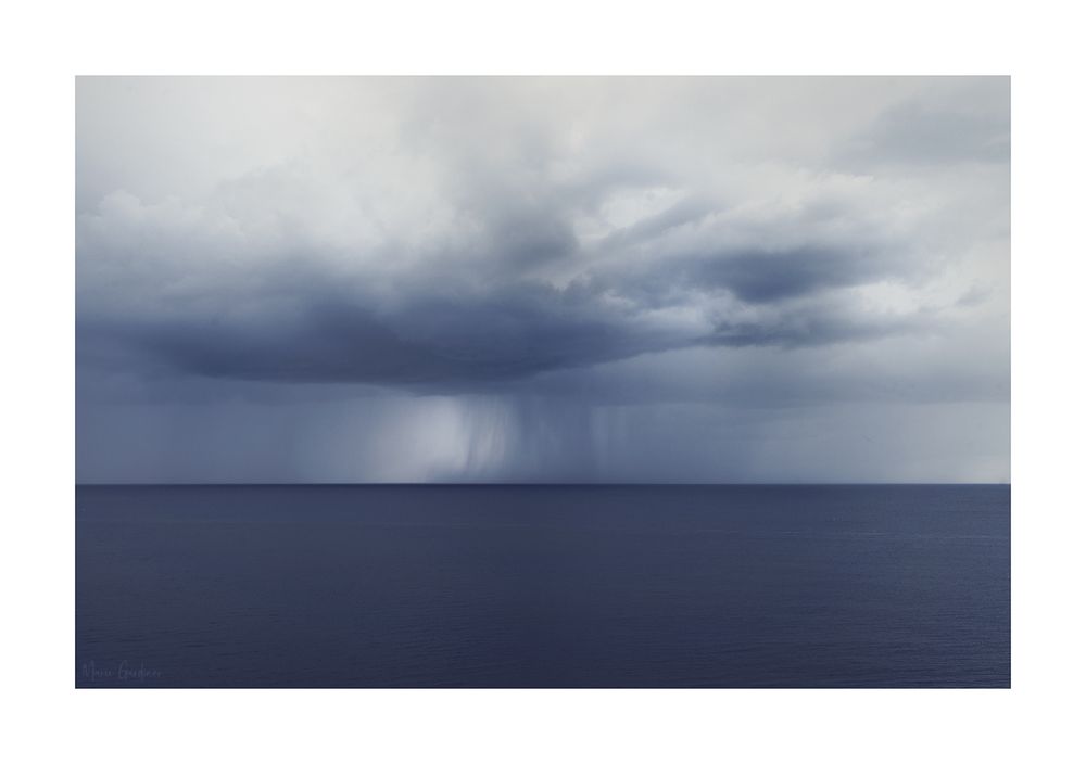 the straight, flat line of the sea (which is very still and calm) but then there's a big storm cloud over it with rain coming down in the distance. The colour of the image is purple-grey-blue