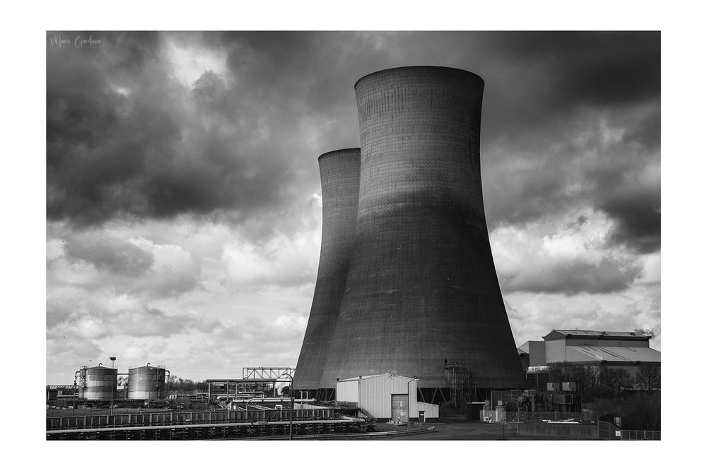 two large cooling towers in a black and white image, with stormy skies above. Various other industrial structures sit below them but pale into insignificance with the size of the towers
