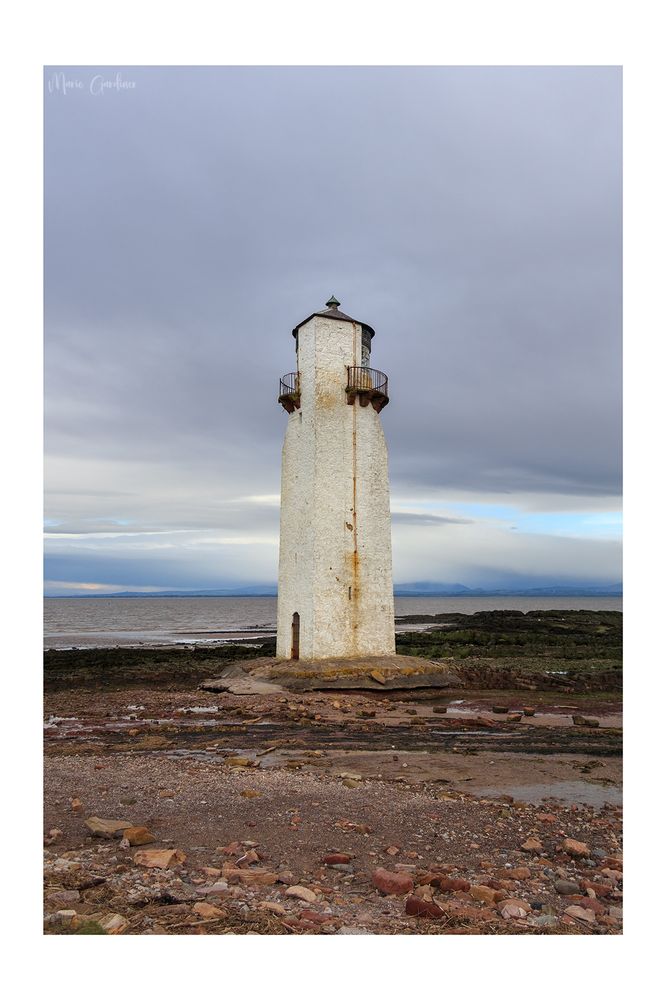 A squareish white lighthouse on rocks with rust and dirt all over. There's a balcony around the top but mostly on the other side of us. In the background is the sea, and the sky is pretty grey and flat
