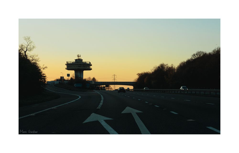 a large concrete tower shaped like an air traffic control tower in the distance, almost silhouetted in the dying light. The motorway (and exit) is in the foreground, with arrows pointing to where to exit.