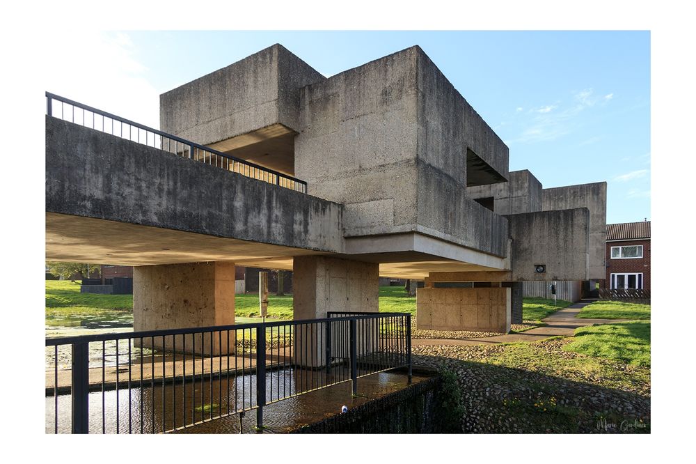 the pavilion from the outside: a mass of concrete block shapes above water with a housing estate behind