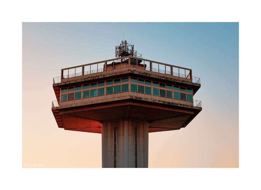 a large concrete tower shaped like an air traffic control tower isolated against a sky that seems to gradient from pale blue to peach 