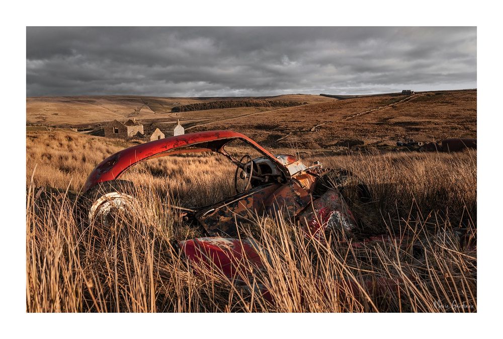 a mostly hidden red shell of a car by grass, there's a ruined house in the background and bare hills behind that 