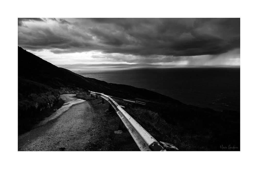 black and white photo of a road and road barrier curving away into the distance down a hill. In the background is the sea, with heavy clouds above and rain coming down in places