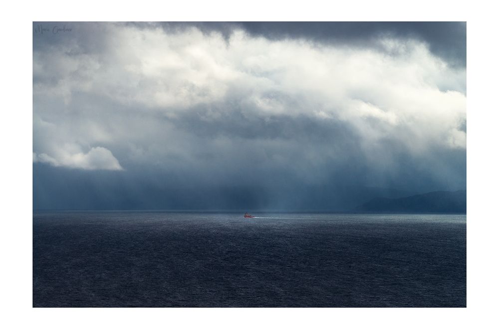 a colour photo of mostly blue hues, with the sea in the foreground and lots of dramatic clouds above with rain coming down in multiple places. In the middle is a tiny orange boat.