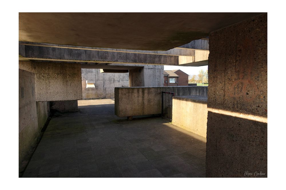the inside of the pavilion, all concrete blocks and shadow and light. There is a graffiti penis just about visible on the wall to the right, so we will see who reads the alt text