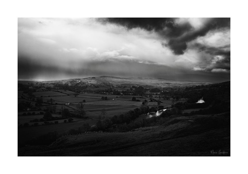 a black and white photo of the hilly countryside with low clouds and rain visible in the near-distance