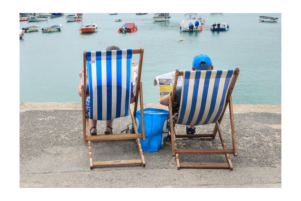 2 blue and white striped deckchairs facing away from us with a couple in them who we can just make out are both reading newspapers. A harbour is behind them