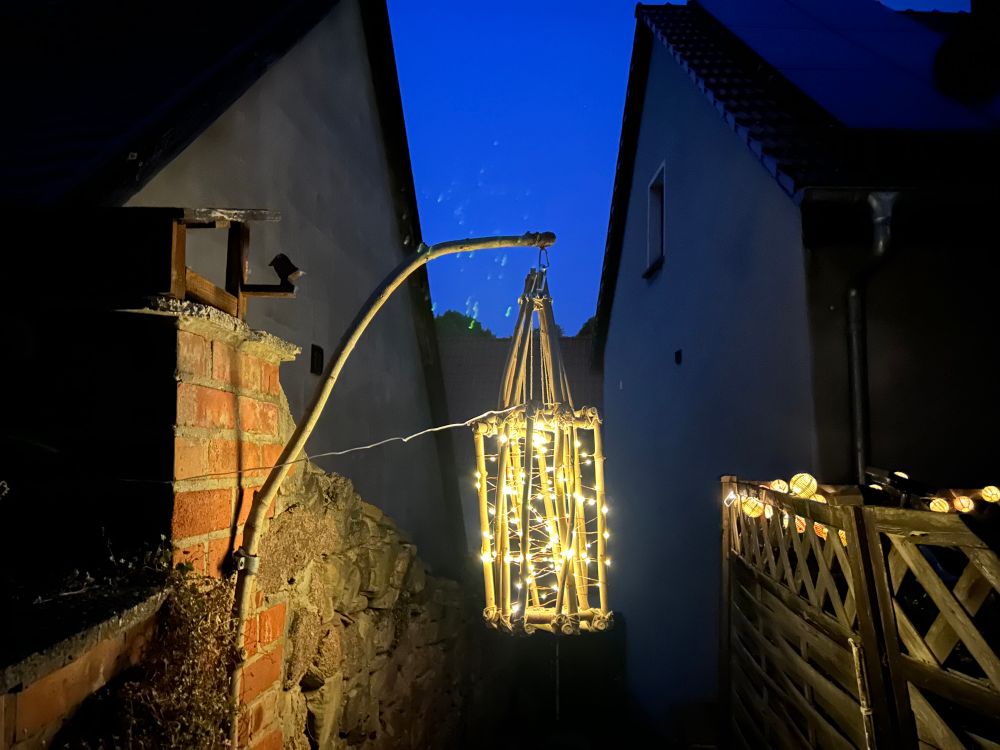 Eine Laterne aus Zweigen gebaut, mit einer Solarlichterkette, die durch die Zweige gewoben wurde. Sie leuchtet in der Dunkelheit nahe an einer Natursteinmauer