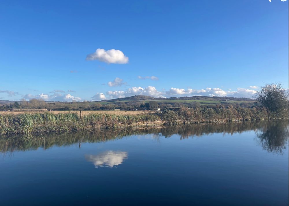 landscape with canal in foreground, green fields and rolling hill side under blue skies with a few white clouds