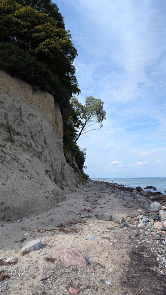 Blick entlang des malerischen Strands und der Steilküste von unten. Der Himmel ist hellblau mit weissen Wolken 