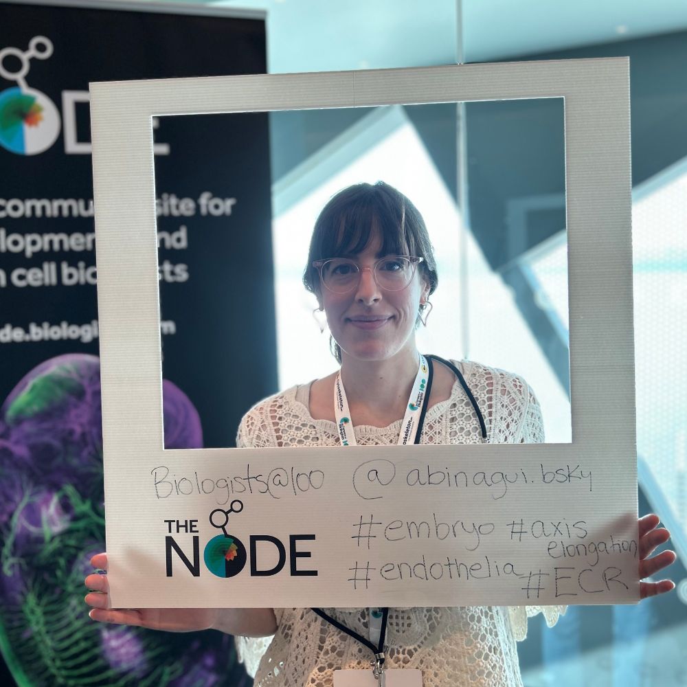 Anahi Binagui-Casas posing with the Node's polaroid frame in front of a roller banner and window. On the frame the text reads "Anahi Binagui-Casas #embryo #axiselongation #endothelia #ECR"