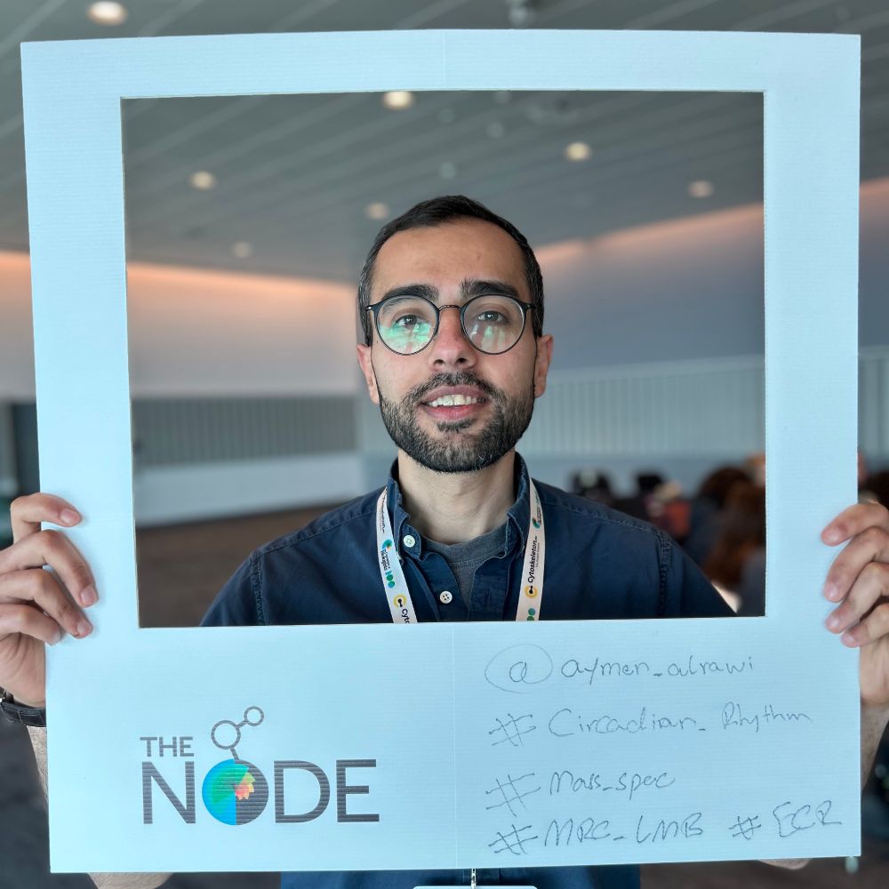 Aymen al-Rawi posing with the Node's polaroid frame. The text on the frame reads "#ECR #CircadianRhythm #MassSpec #MRC_LMB"