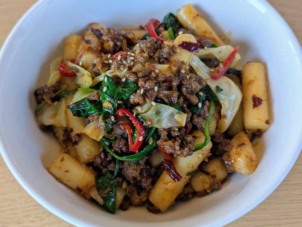 A bowl of spicy bulgogi beef with wilted greens, cabbage, sliced chili, toasted sesame seeds, gochujang chili flakes and rice cakes