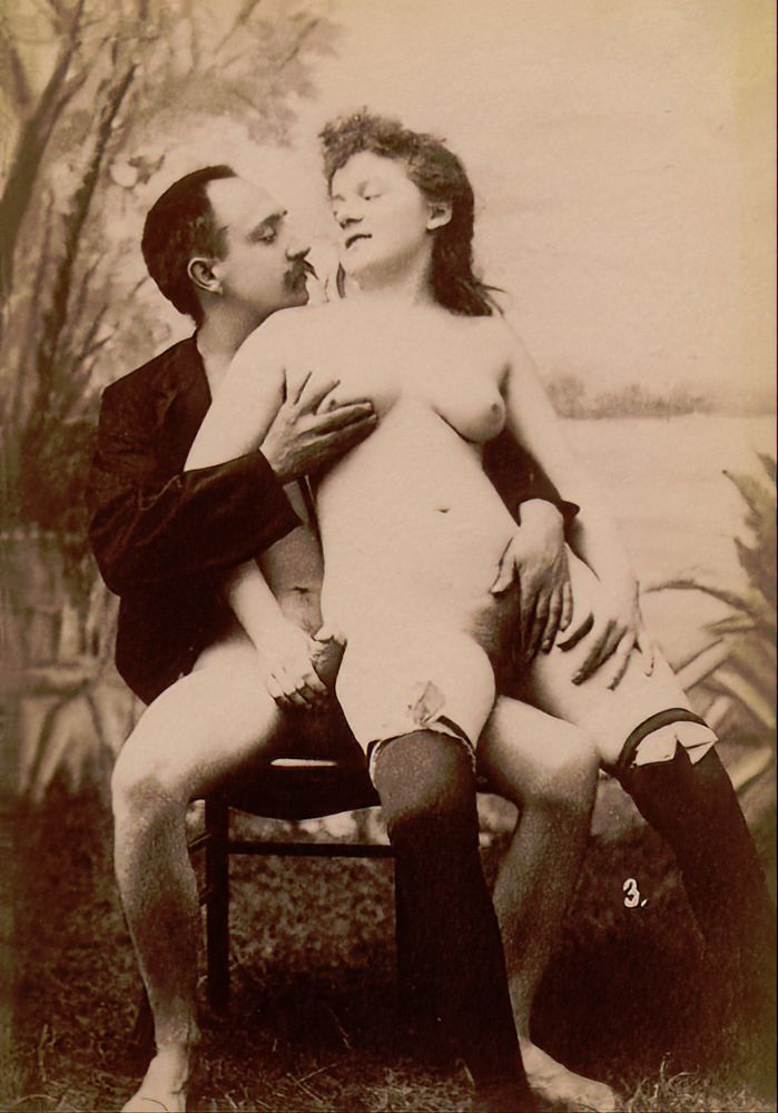 Early 1900s sepia photo. Man sitting on a chair. He has a large mustache. He is wearing a jacket, but nothing else. On his knee, facing us, is a long-haired woman, naked except for black stockings held up just above the knees with white bows. The man holds the woman's right breast. The woman holds the man's erect penis. They look in each other's face.