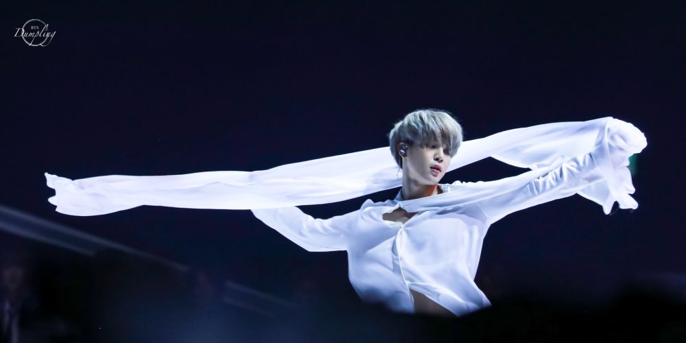 Jimin is dressed all in white with a flowing scarf extended horizontally behind him as he dances to the orchestral arrangement of "I Need U." His hair is silvery.