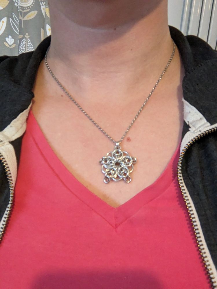 A star shaped necklace made from bright aluminium jump rings. It is worn on a feminine body, showing the chest and torso, the person is wearing a pink v-neck t-shirt and black zip top