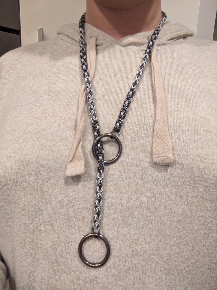 A single length of Byzantine weave chainmaille chain, in alternating black and white segments, capped with steel rings. The chain is woven through one of the big rings, making it a closed piece. It is worn on a chest covered in a cream top