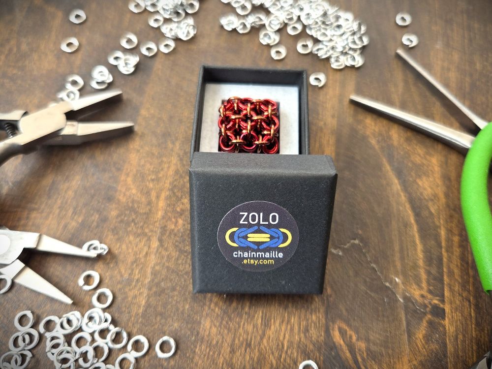 A red and brown mini fidget cube in a black jewellery box with an offset lid with my logo on it (ZOLO over a stylised Byzantine link in blue and yellow). The box is on a dark brown wooden surface, surrounded by silver jump rings and green-handled pliers.