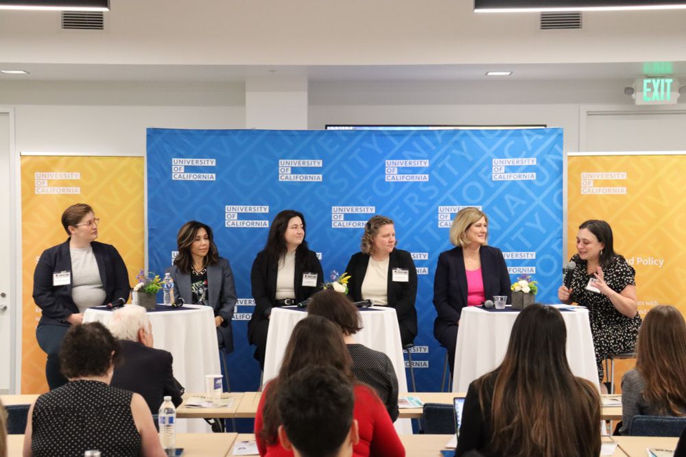 6 white women in suits on a panel with a blue and yellow UC step and repeat background