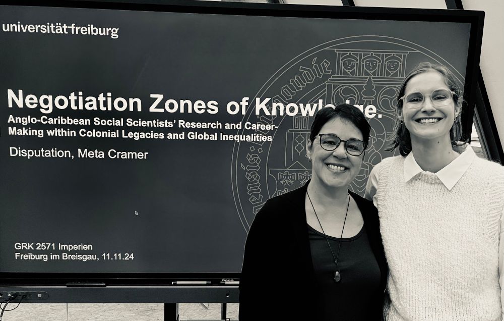 two persons standing smiling in front of a large screen with the dissertation titel displayed.