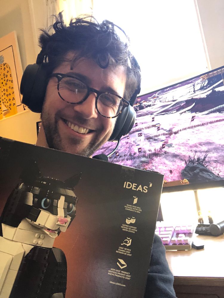 A photo of James hold a tuxedo cat Lego set in front of a computer monitor displaying a battle in Total War: Warhammer. 