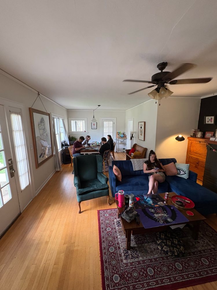 Four people around a dinner table playing magic, and a coffee table and couches around another in-progress game of magic. 