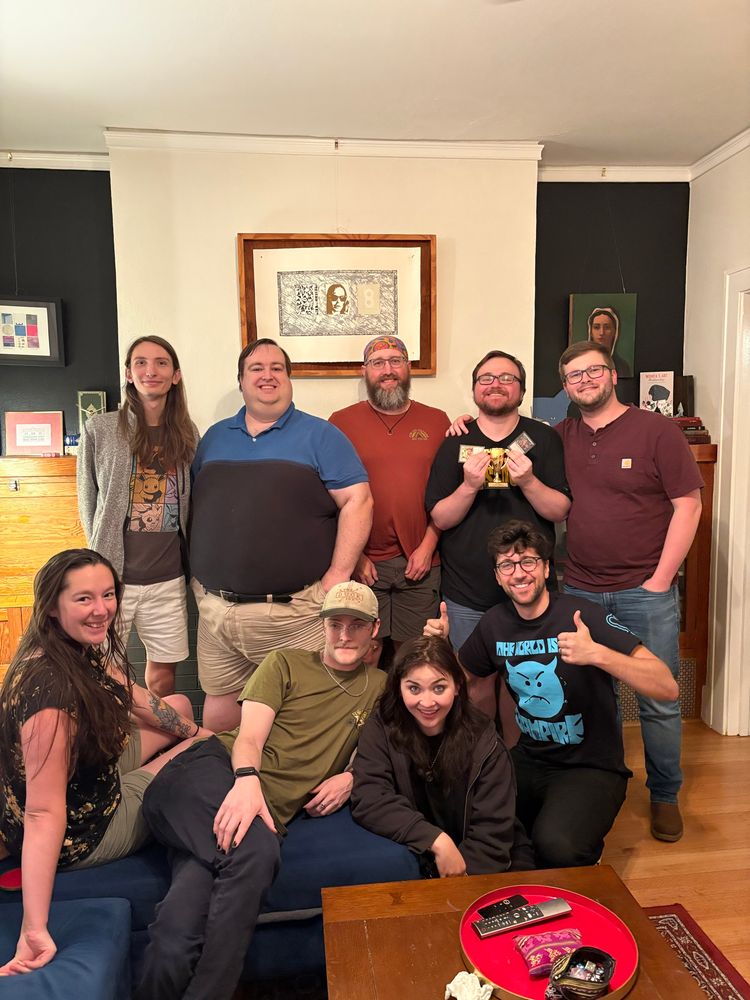 Nine people posing for a photo. One holds a trophy, some hold magic cards. 