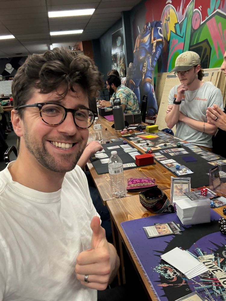A photo of me giving a thumbs up at a table with magic the gathering cards. 