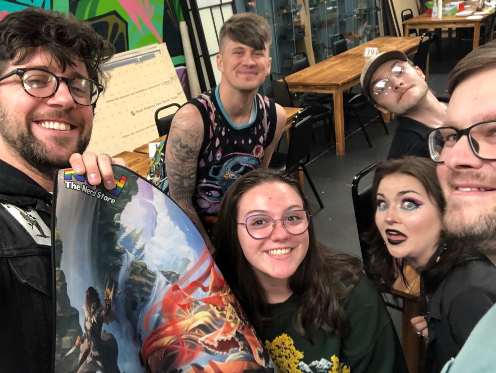 Me holding up the playmat I won at a magic the gathering prerelease and smiling with the five friends that posed for the selfie photo with me. 