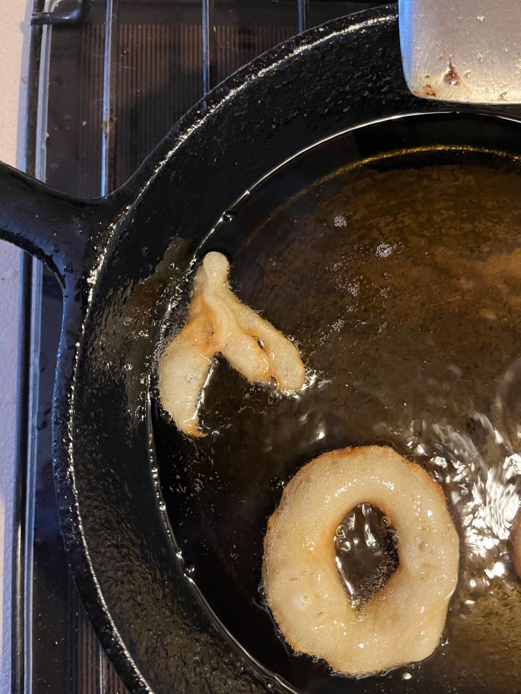 Two homemade doughnuts frying in a cast iron fry pan, one is misshapen and resembles a clitoris