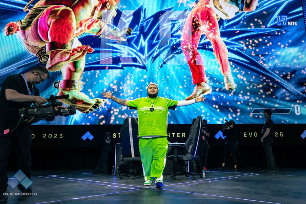 Photo of MenaRD reacting to winning Street Fighter 6. He has turned to face the crowd, arms outstretched. The arena's LED screen is behind him with the game's "KO" graphics still showing.