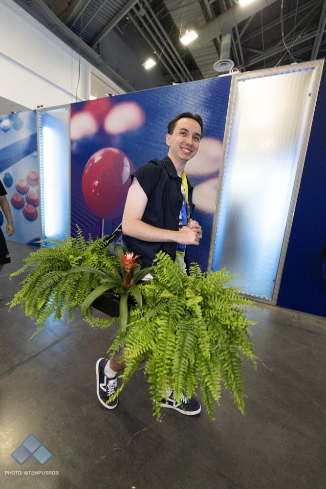 A man carrying a planter, with large ferns growing in it. The planter also contains a fully functioning arcade stick.